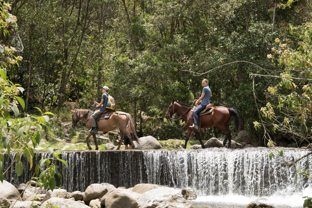 Zwei Reiter reiten über einen Fluss mit einem kleinen Wasserfall im Vordergrund