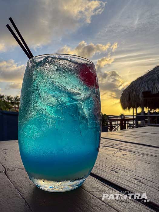 Longdrinkglas auf einem dunklen Holztisch vor dem Sonnenuntergang mit eisgekühltem blauem Curacao Likör, roter Cocktailkirsch und zwei Strohalmen