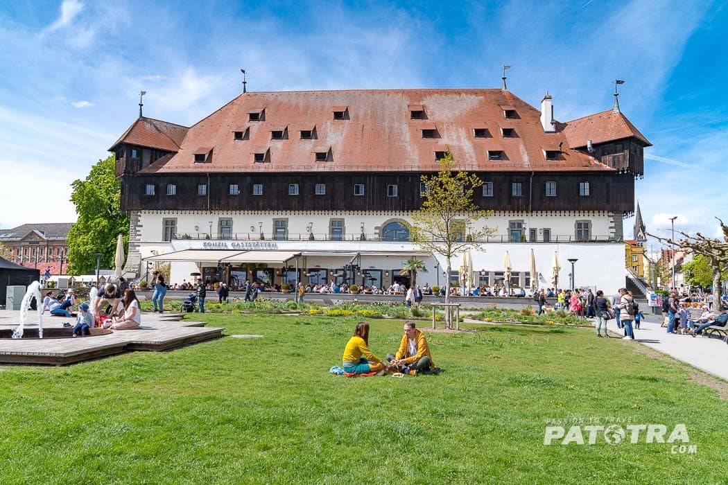 Das Konzilgebäude vom See aus mit zwei Personen sitzend im Rasen davor